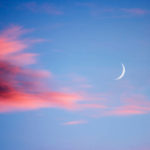 New moon at sunset with light clouds in various shades of red and magenta, darker to the left and a blue sky behind.